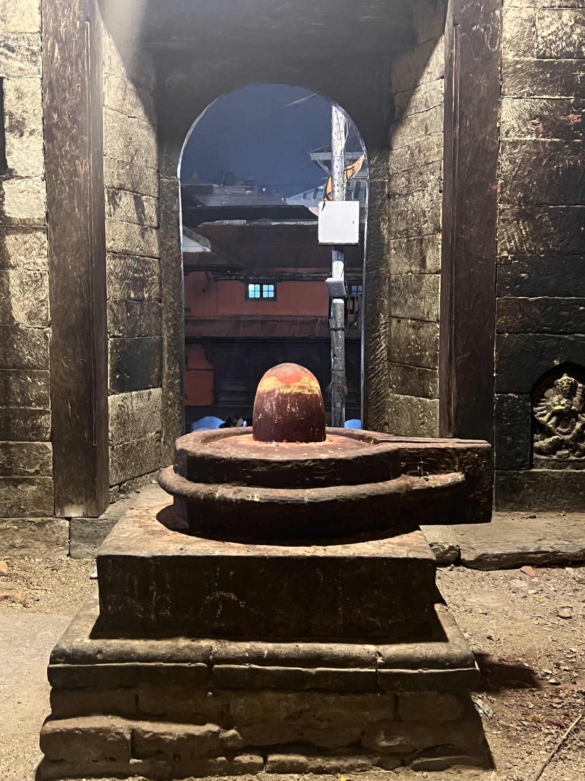 A stone Shivalinga shrine framed by a dark stone archway, glowing softly at night