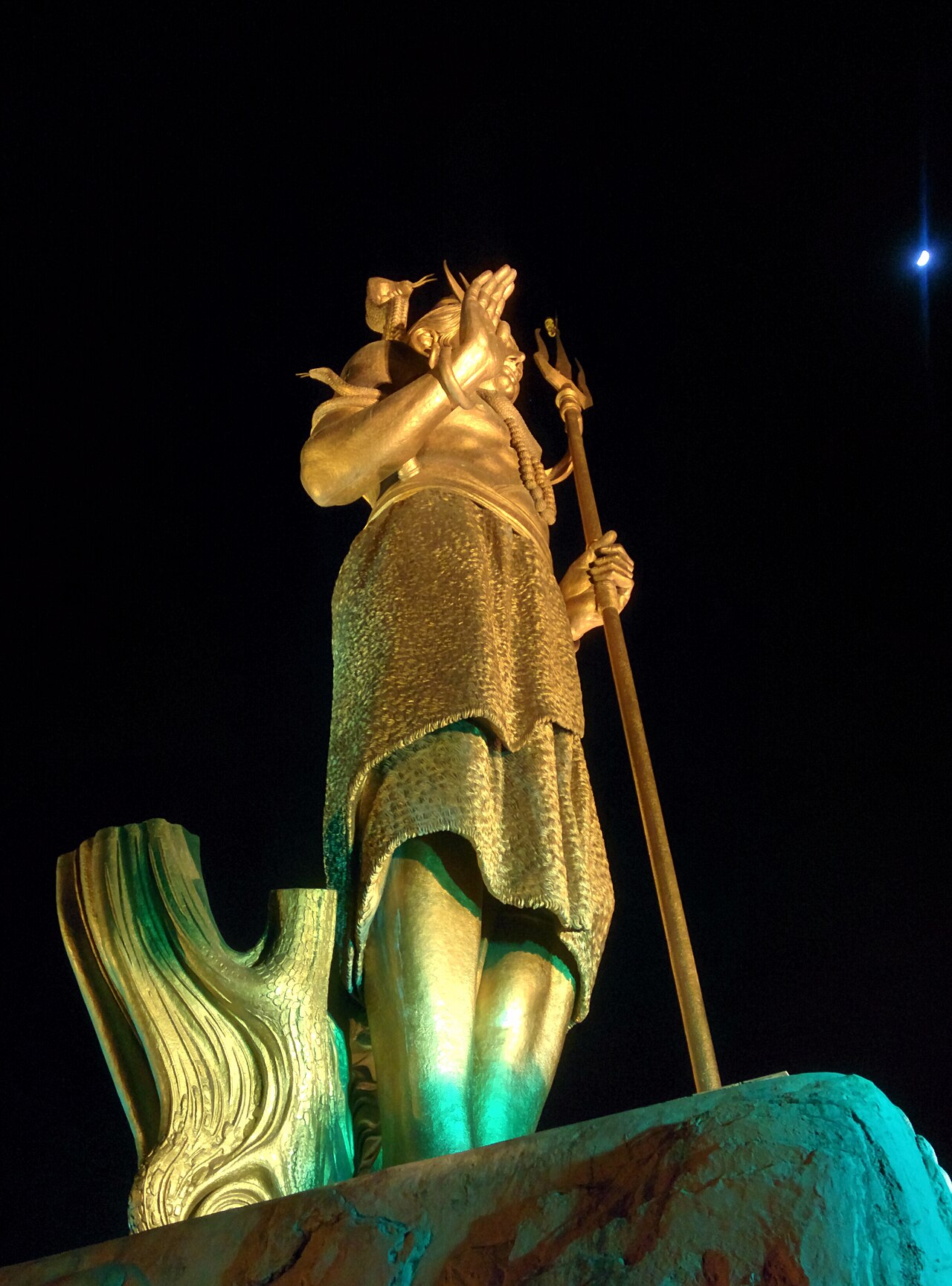 Golden statue of Lord Shiva with trishul at night, the moon visible in the dark sky