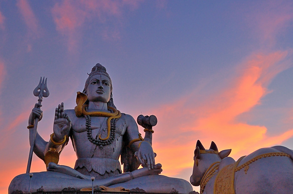 The Murudeshwar Shiva statue against a pink and violet sunset sky, with Nandi the bull beside him